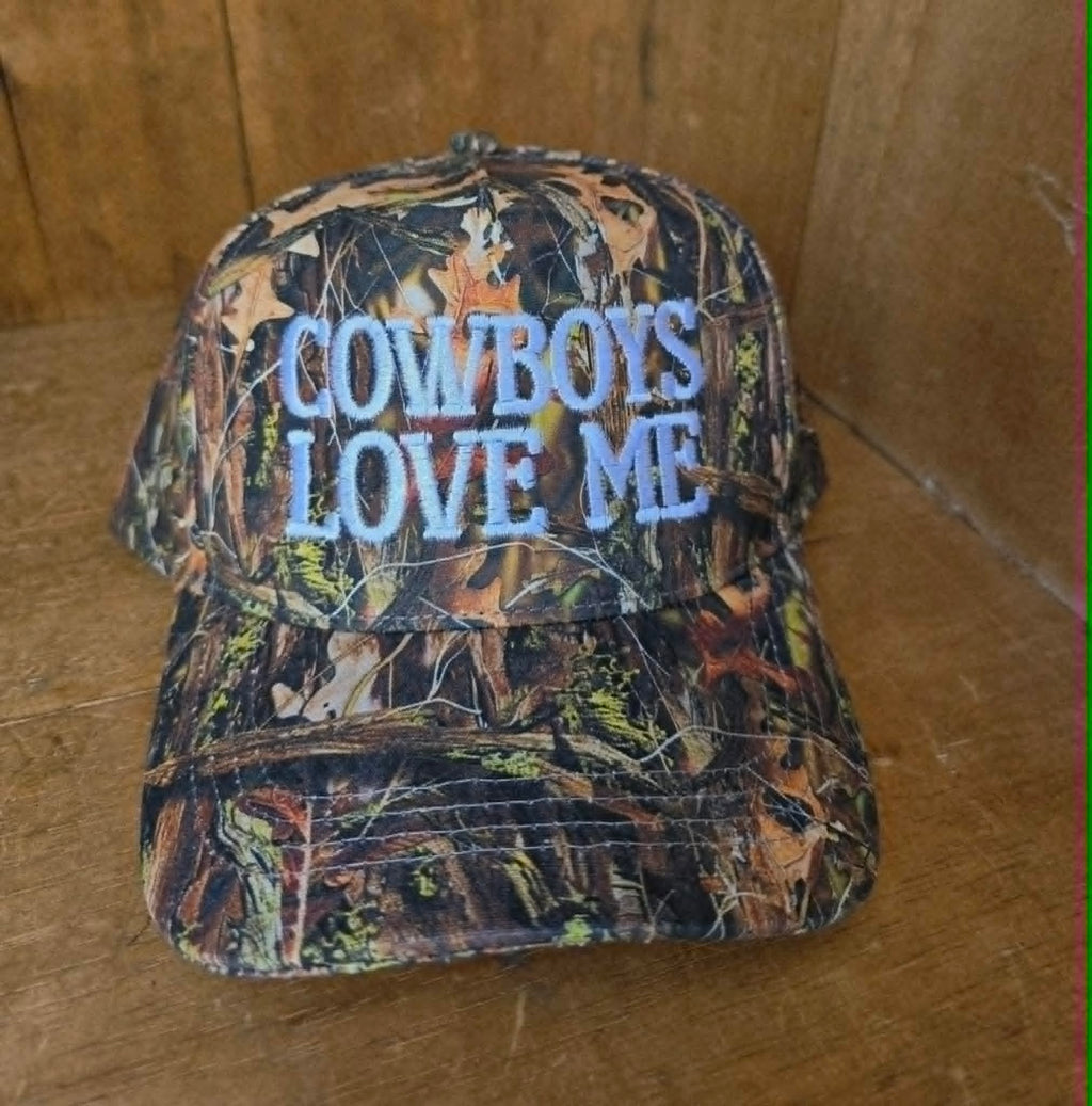 Dusty Cactus Co camo trucker cap with statement "Cowboys Love Me" embroidery, styled on model with neutral western jacket for an edgy country inspired outfit