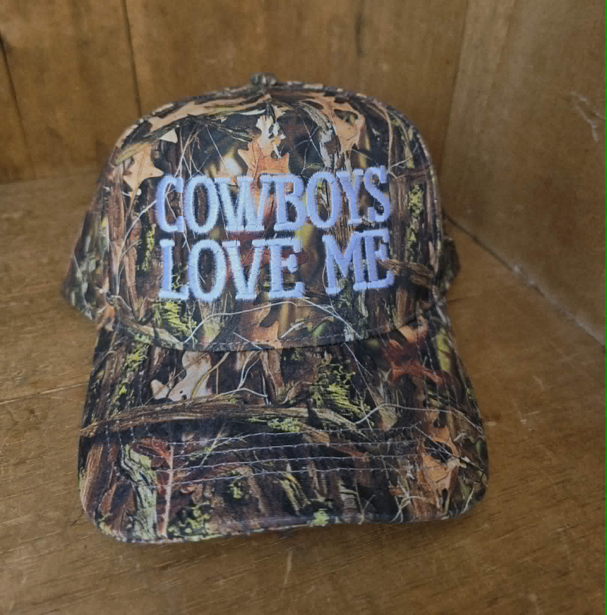 Dusty Cactus Co camo trucker cap with statement "Cowboys Love Me" embroidery, styled on model with neutral western jacket for an edgy country inspired outfit
