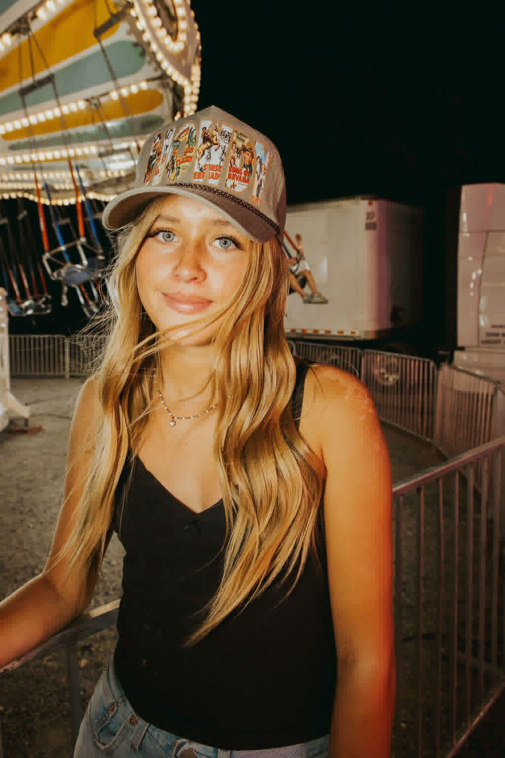 Beige trucker cap with a structured front panel featuring bold block lettering filled with a collage of vintage Western imagery, including cowboys, rodeo scenes, and retro posters. The brim is slightly darker beige with a worn, textured finish, and a brown rope detail runs along the base of the front panel for a classic Western touch.