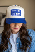 Western trucker cap in cream and royal blue with curved brim, featuring bold patch that reads "Cowgirl at Heart". 