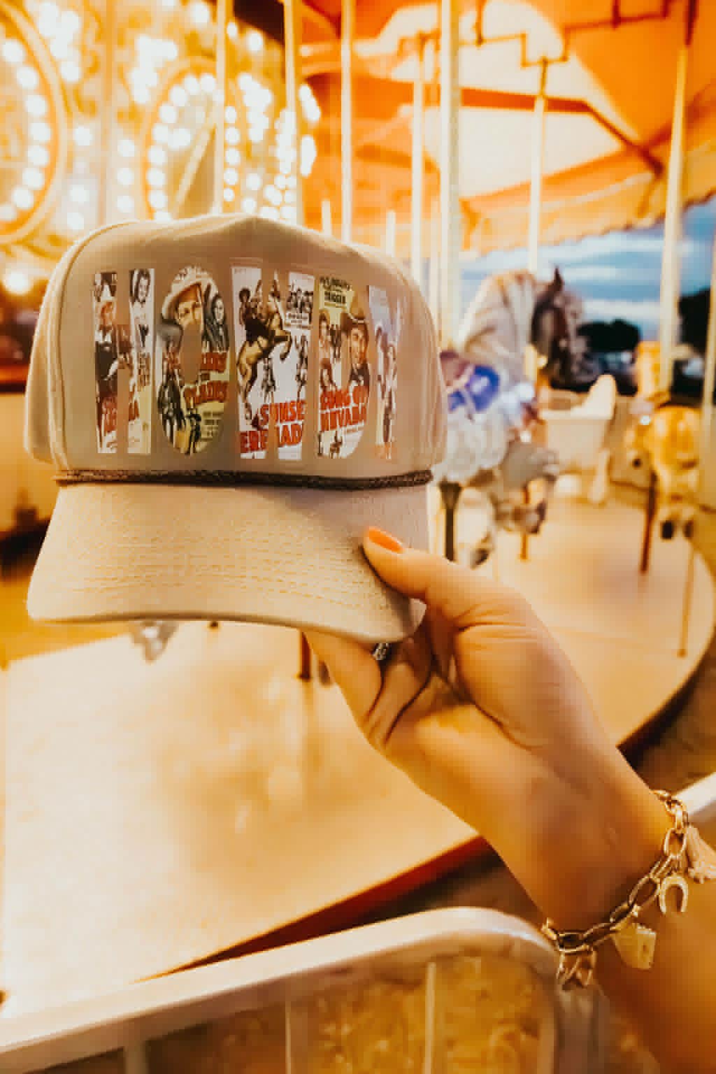 A hand holds a beige trucker cap in front of a brightly lit carousel. The cap features a collage-style graphic across the front panel with vintage Western-themed images forming bold lettering. The brim is a slightly darker beige with a textured finish. In the background, carousel horses and warm golden lights create a nostalgic, carnival-like atmosphere. The person holding the cap wears a gold bracelet with small charms, adding to the styled, Western-inspired aesthetic.