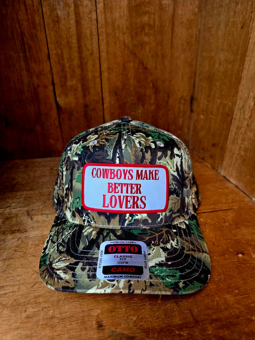 Camo western trucker cap with bold red and white "Cowboys Make Better Lovers" patch, styled outdoors with a country inspired aesthetic