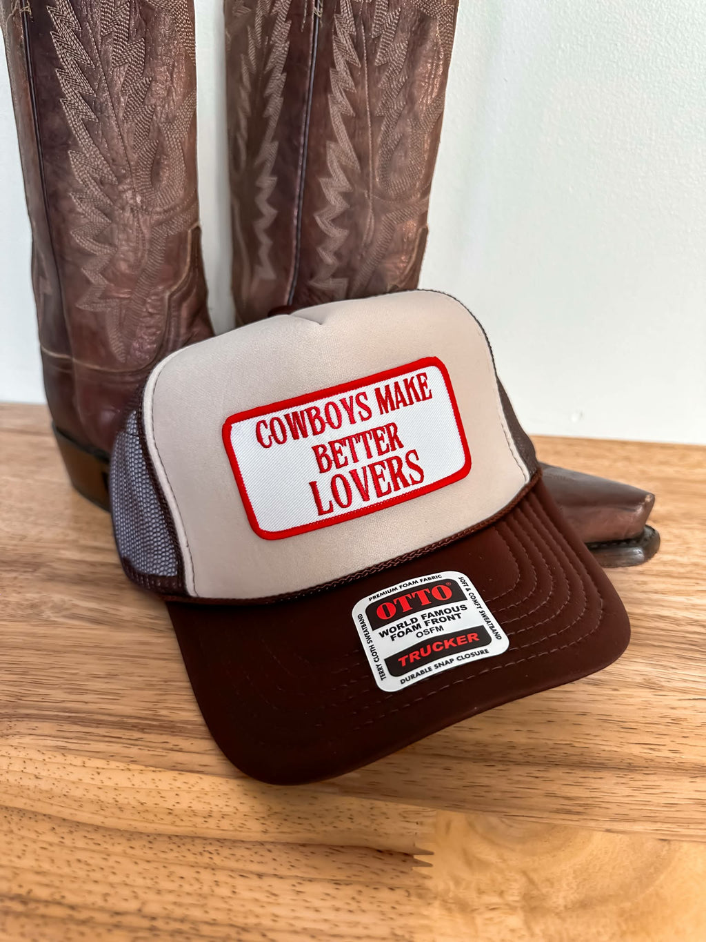 Two toned western trucker cap in beige and brown with a mesh back and curved brim, showcasing a statement patch that says "Cowboys Make Better Lovers" in red lettering.