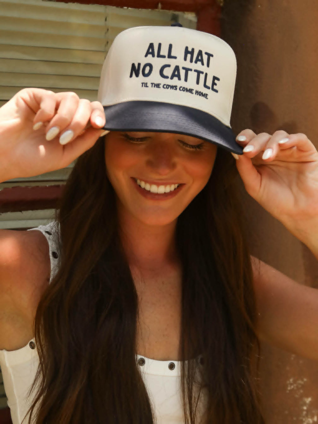 Women wearing a navy and cream western trucker cap embroidered with "All Hat No Cattle Till The Cows Come Home" while holding the brim outdoors in warm sunlight