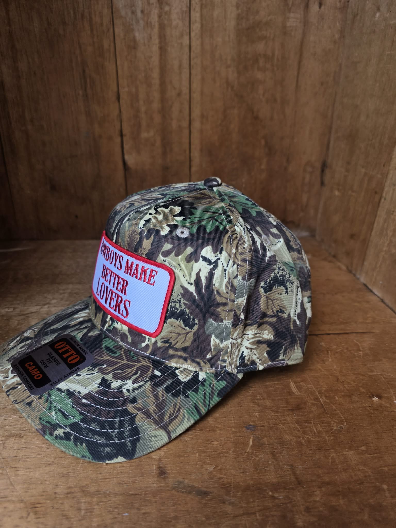 Dusty Cactus Co Camouflage trucker cap featuring embroidered "Cowboys Make Better Lovers" patch on a structured curved brim cap