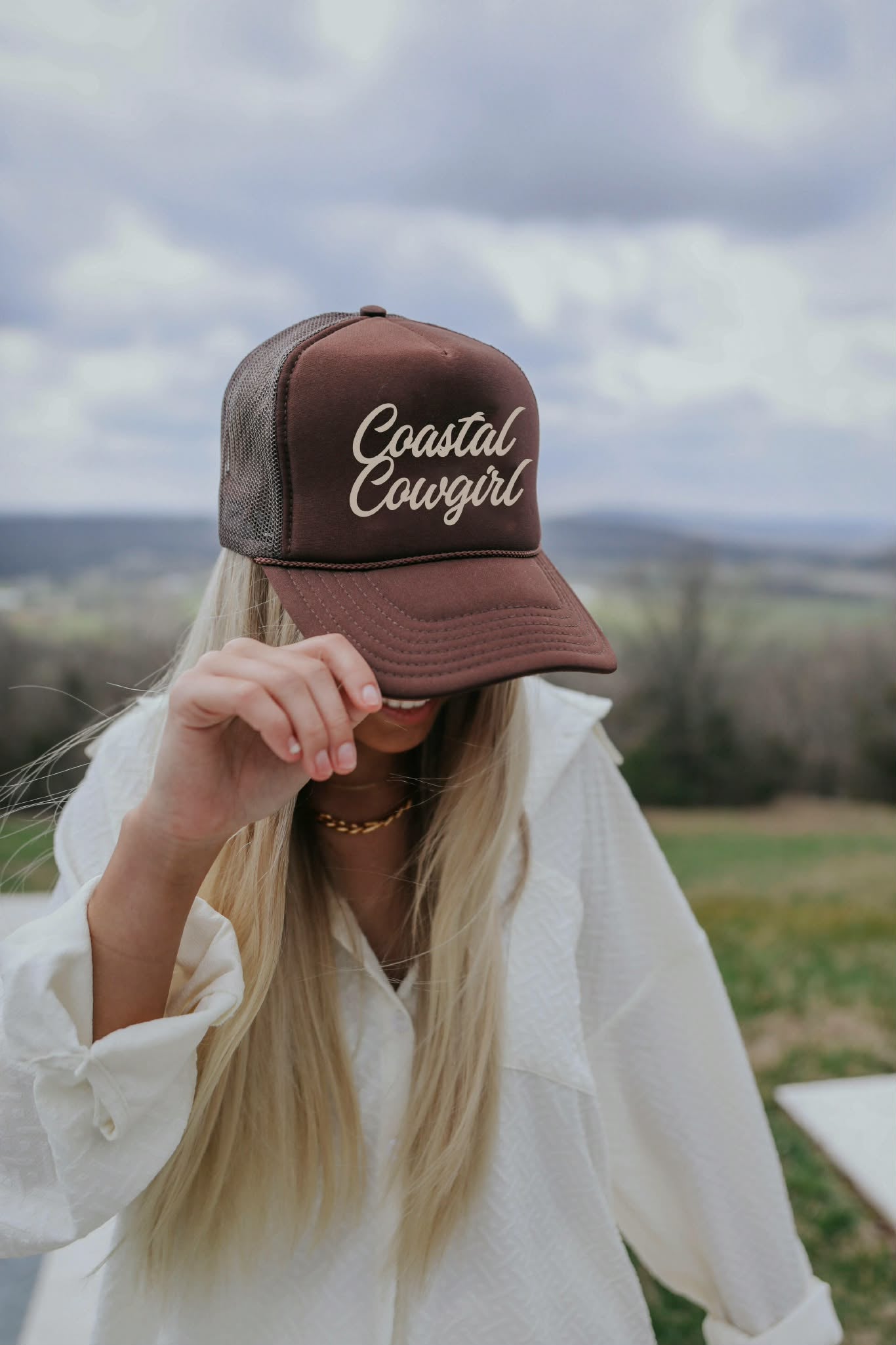 Brown mesh back trucker cap with " Coastal Cowgirl" embroidered in gold script, styled outdoors with soft neutral country aesthetics