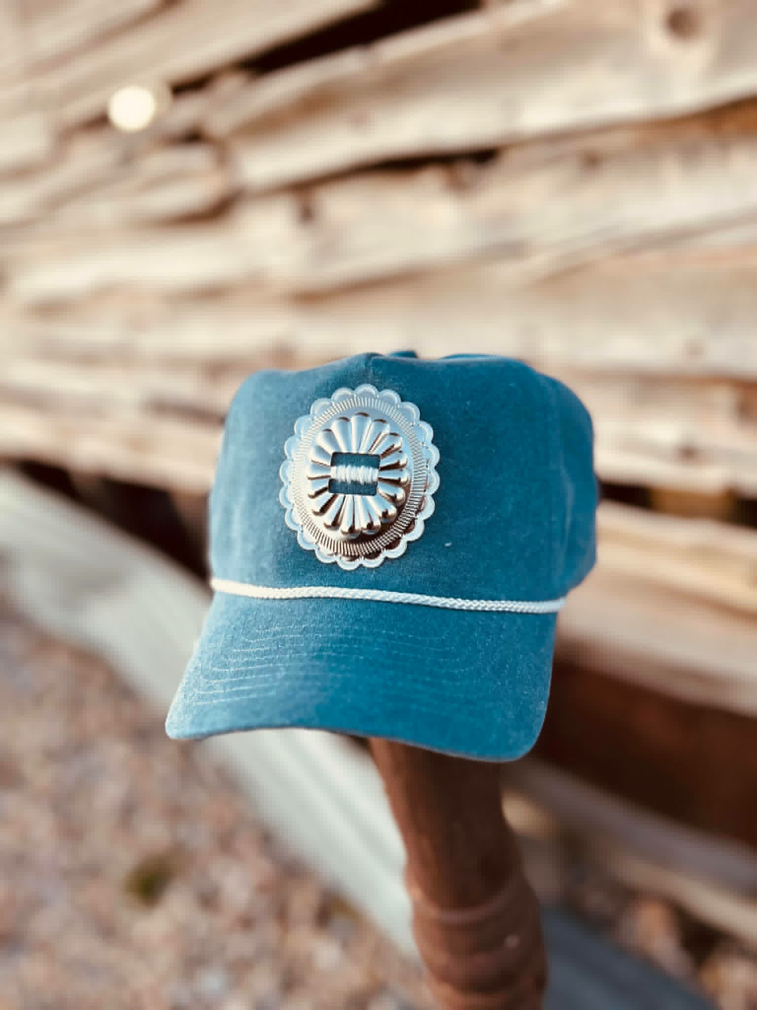 Denim trucker cap in a faded blue wash with a structured front panel featuring a silver concho-style embellishment centered on the front. The cap has a curved brim, a white rope detail across the base of the front panel, and a classic Western-inspired design