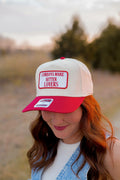 trucker cap with a bold red brim and embroidered patch reading “Cowboys Make Better Lovers.” The western-style cap features a mesh back and red contrast detailing, photographed at sunset with a soft blurred country backdrop.