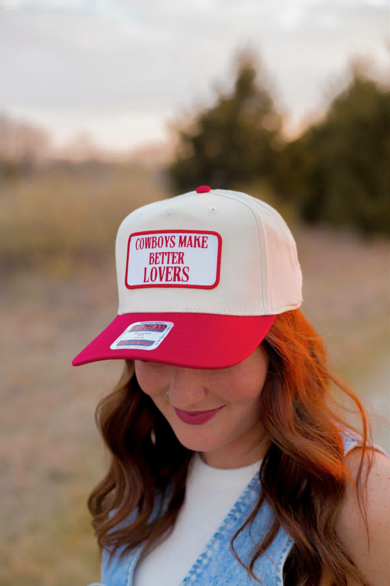 trucker cap with a bold red brim and embroidered patch reading “Cowboys Make Better Lovers.” The western-style cap features a mesh back and red contrast detailing, photographed at sunset with a soft blurred country backdrop.