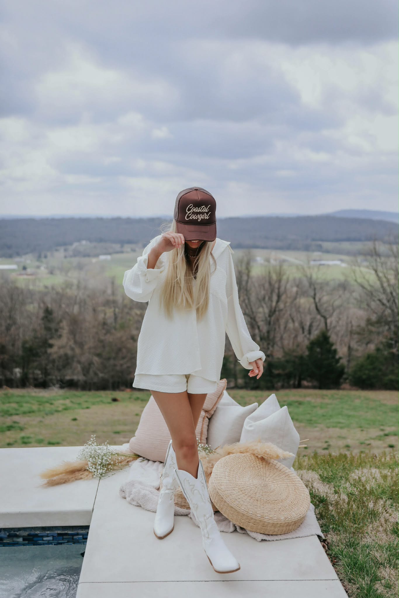 Western inspired Coastal Cowgirl trucker cap in deep brown with curved brim and mesh back, Photographed against a scenic countryside backdrop.