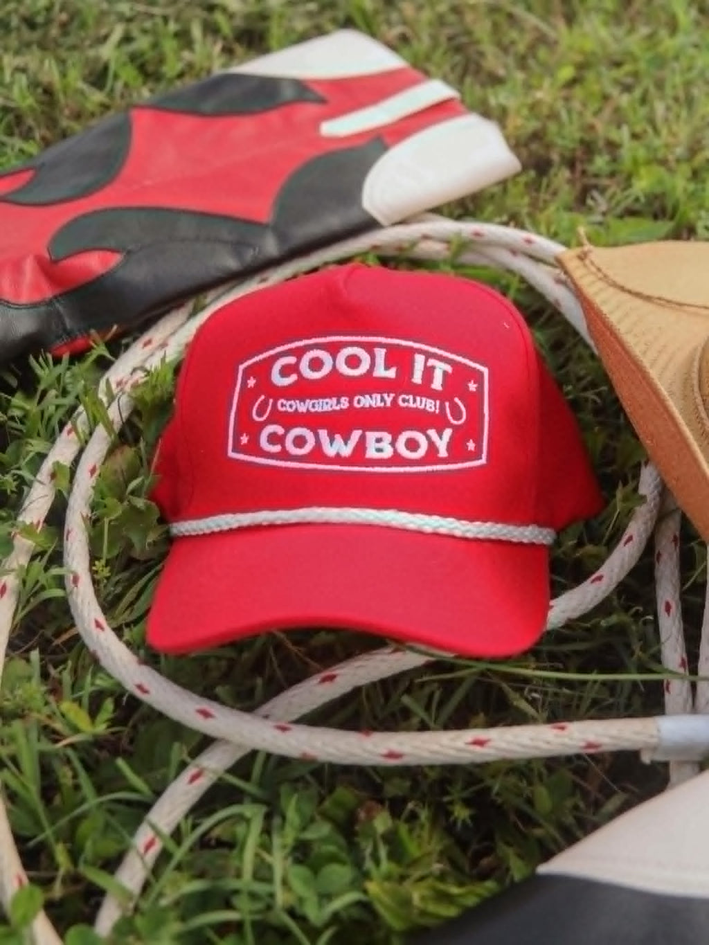 Bright red cap western cap featuring bold white embroidery reading "cool it cowboy" with "Cowgirl only club" detailing, styled with white rope trim and photographed amongst rodeo inspired prop and greenery