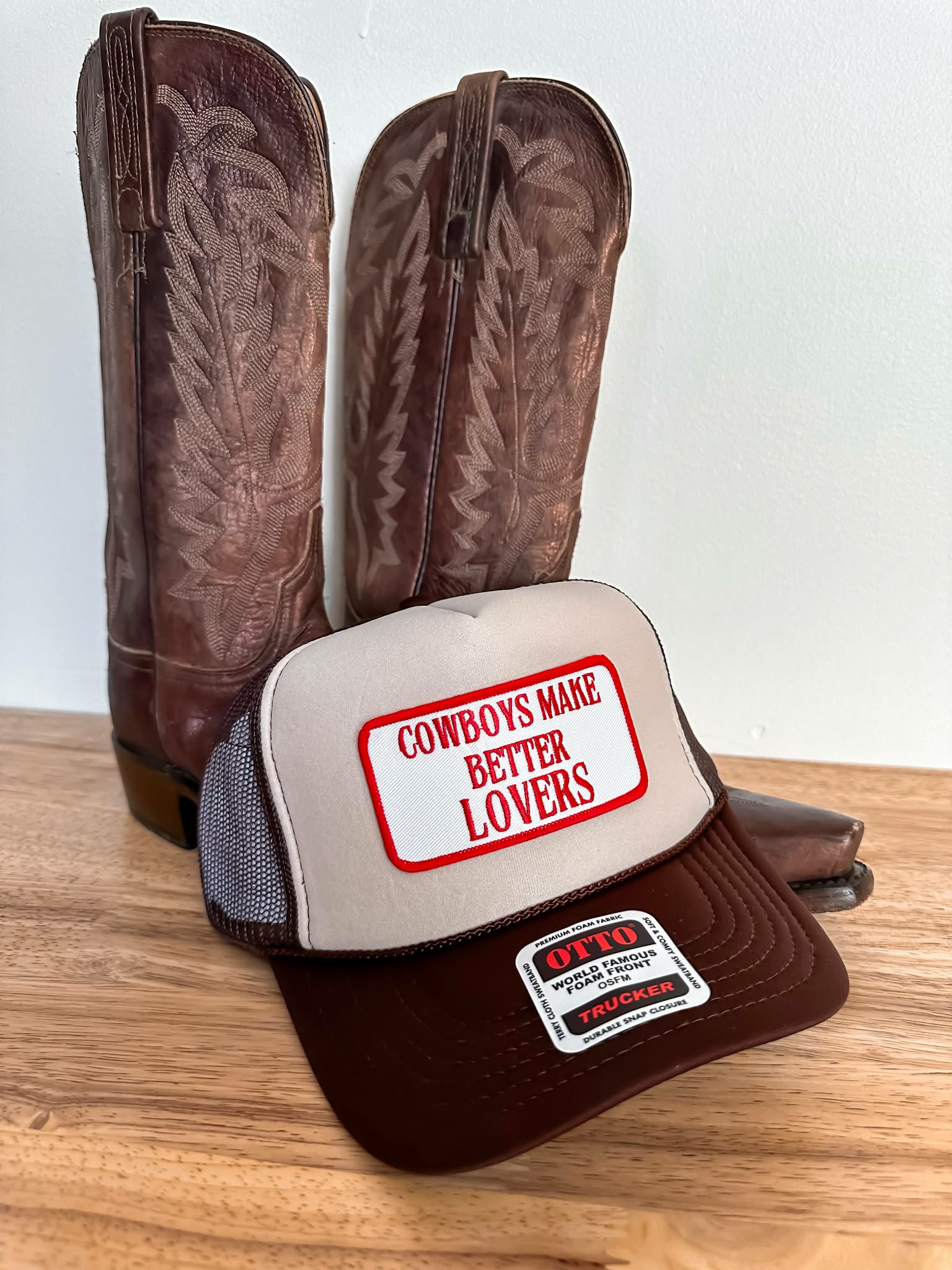 western style trucker cap with cream crown and chocolate brown brim, featuring bold red embroidered text on patch reading "Cowboys Make Better Lovers"