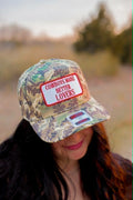 Camouflage trucker cap with an embroidered white patch outlined in red reading “Cowboys Make Better Lovers” in red lettering. The western-style cap features an all-over woodland camo print with a curved brim and structured front panel, 
