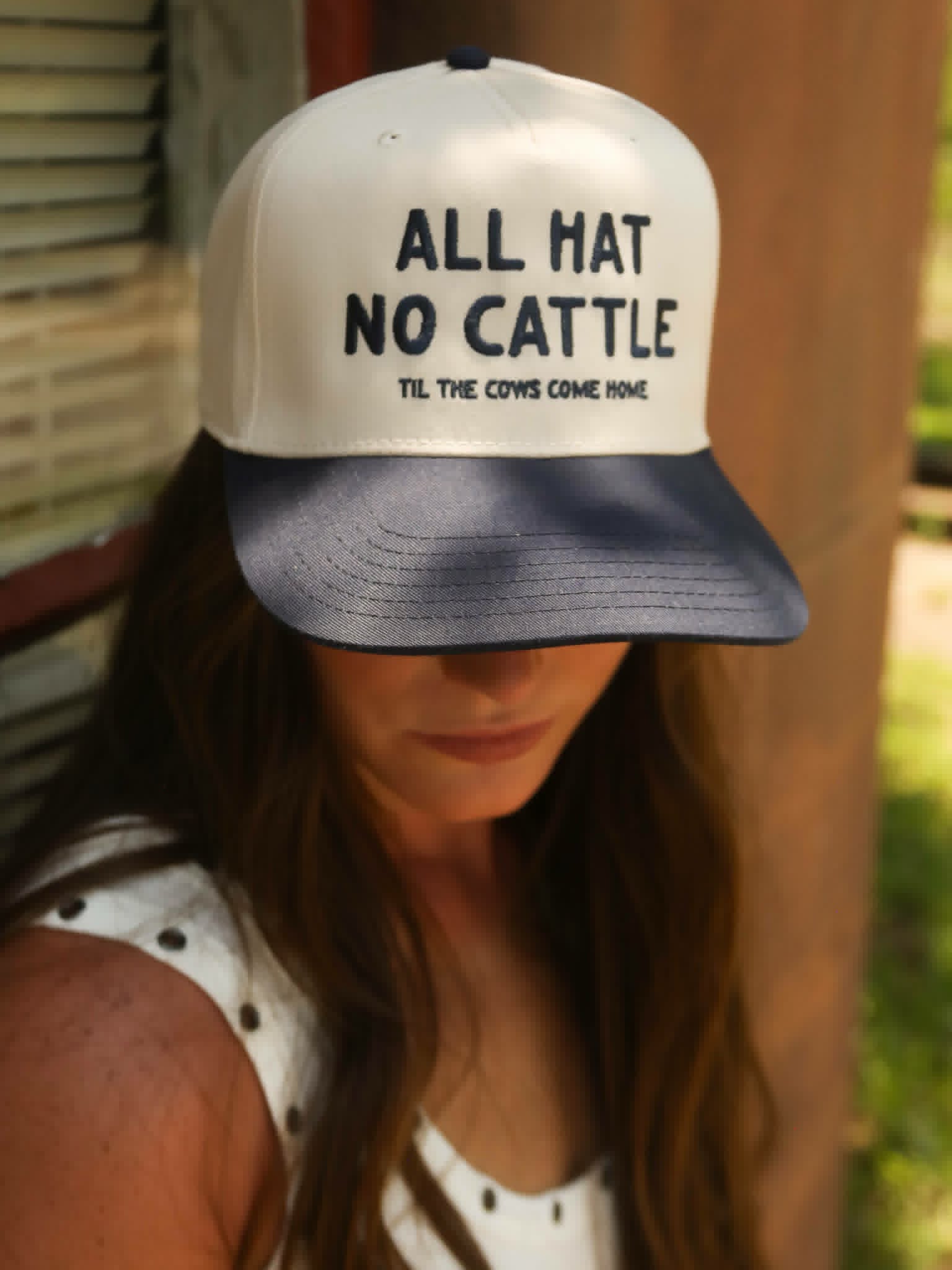 Western inspired navy brim trucker cap its bold embroidered slogan "All Hat No Cattle" worn with a white ribbed top and long brunette hair.