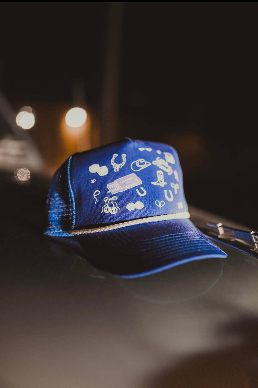 Navy blue trucker cap with a structured front panel featuring scattered white embroidered Western-style icons, including horseshoes, a cowboy hat, dice, boots, and small playful symbols. The cap has a curved brim, a white rope detail across the base of the front panel, and a mesh back, shown resting on a car under soft nighttime lighting