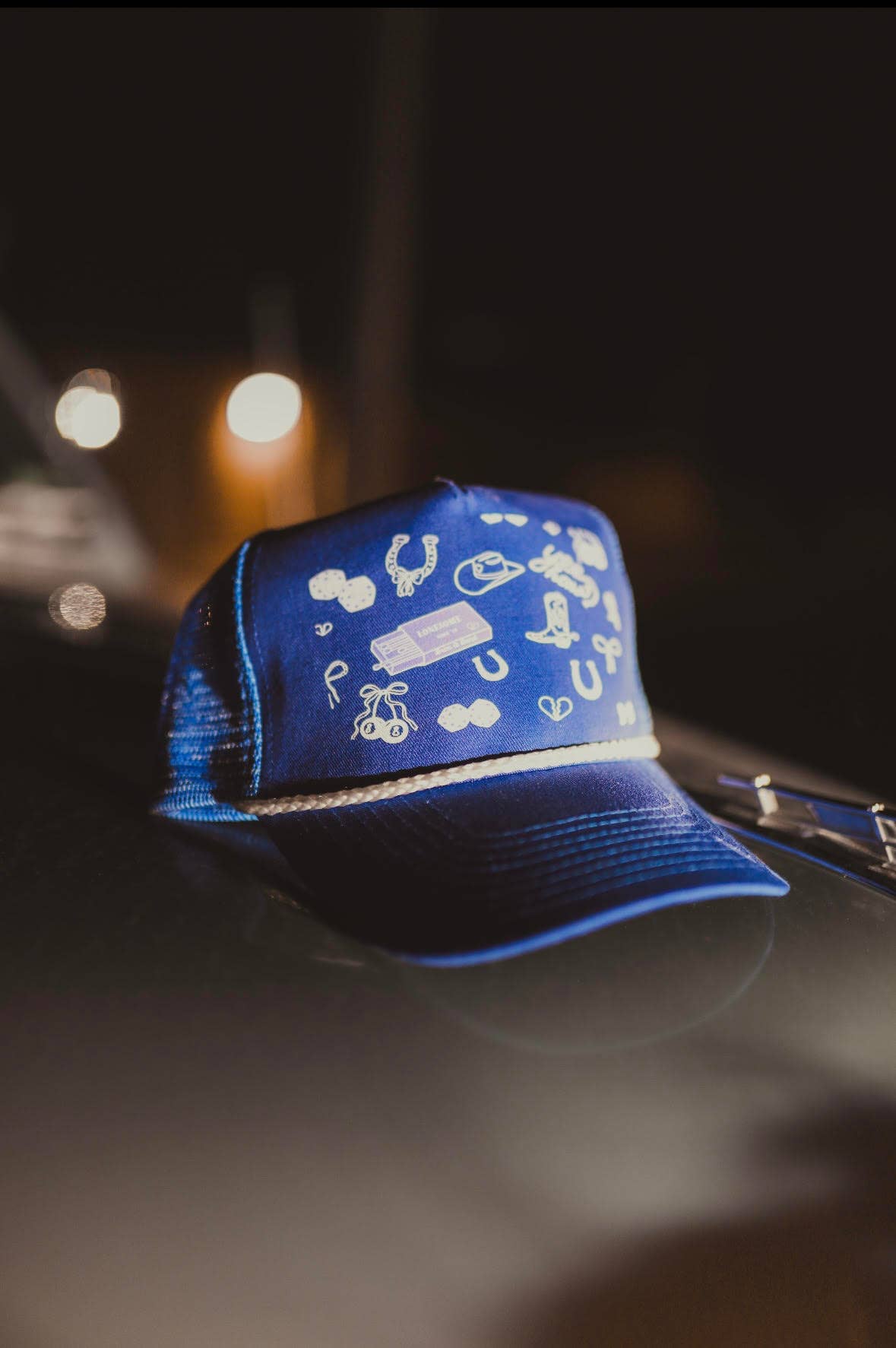 Navy blue trucker cap with a structured front panel featuring scattered white embroidered Western-style icons, including horseshoes, a cowboy hat, dice, boots, and small playful symbols. The cap has a curved brim, a white rope detail across the base of the front panel, and a mesh back, shown resting on a car under soft nighttime lighting